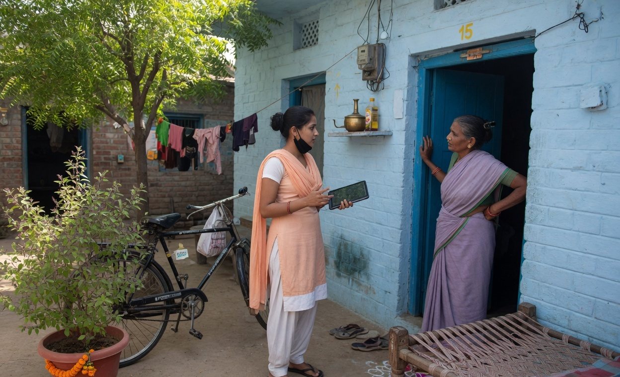 An enumerator with a tablet speaking to a woman at a village doorway in India